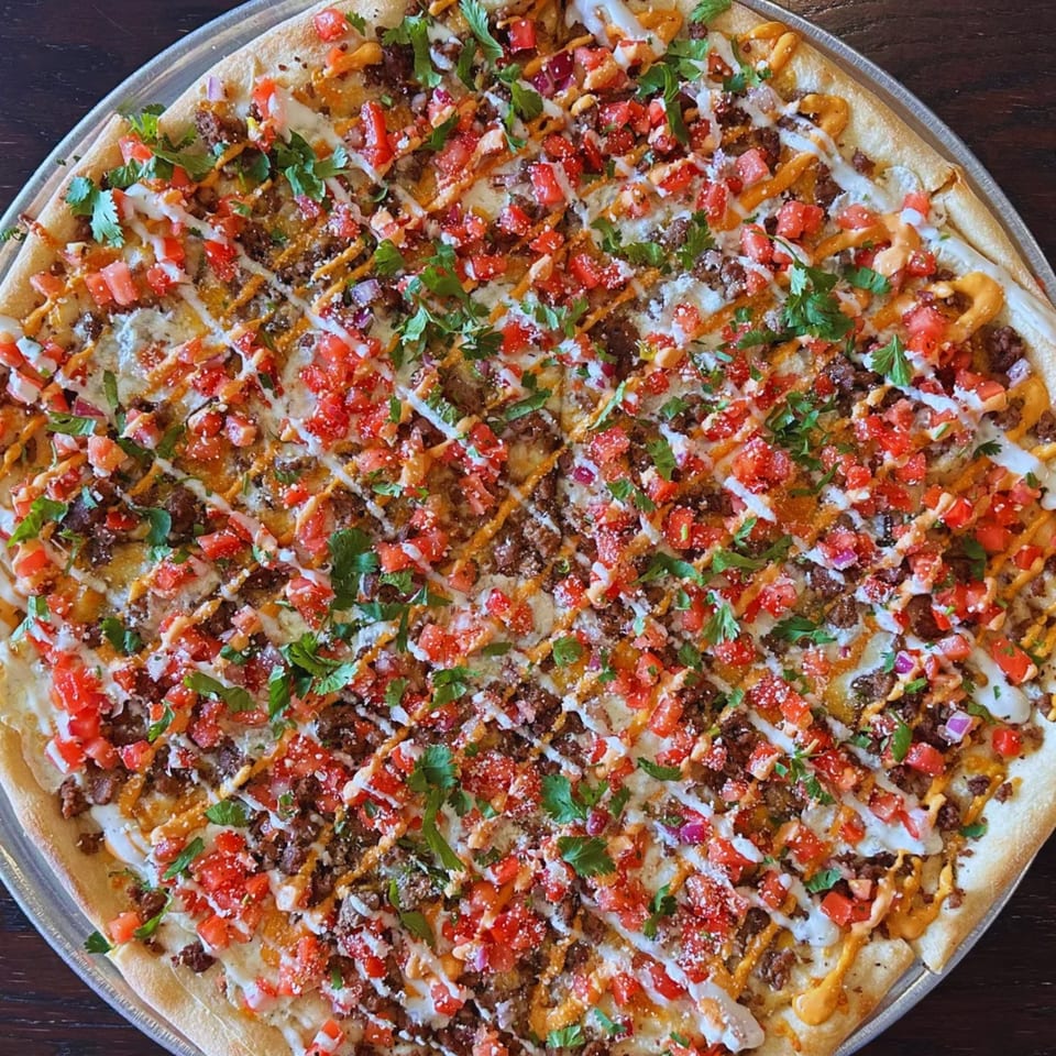 Overhead view of a loaded flatbread topped with seasoned beef, diced tomatoes, onions, parsley, melted cheese, and drizzled sauces.