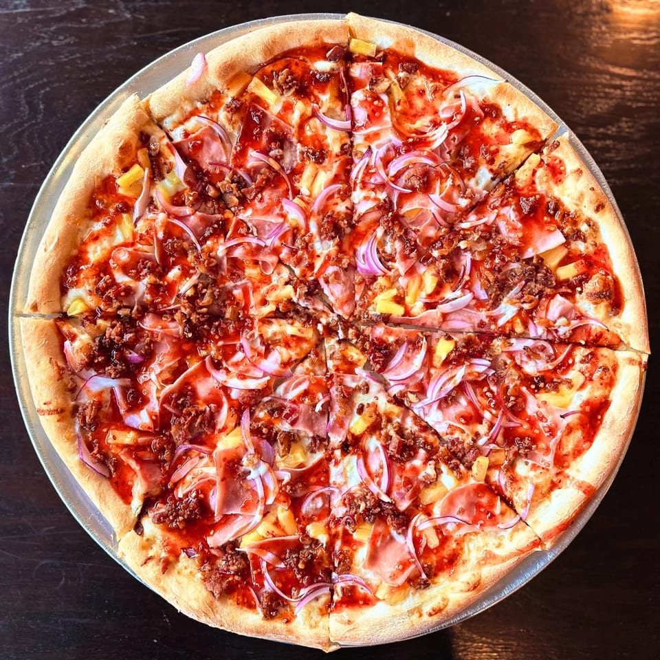 Large sliced pizza topped with tomato sauce, cheese, ham, ground meat, pineapple, and red onions on a metal tray.